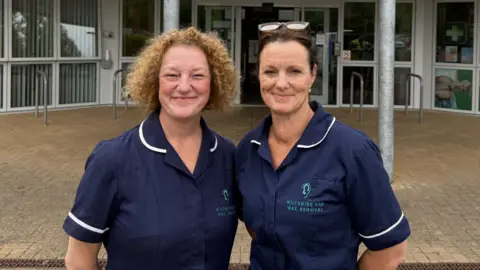 Two women in blue nurse-style uniforms stand outside a building and face the camera