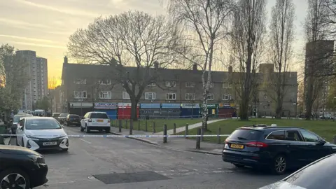 A suburban street with low rise buildings and cars parked on both sides of the road. There are trees in and green hedges and grass. A block of flats is visible at the end of the street.