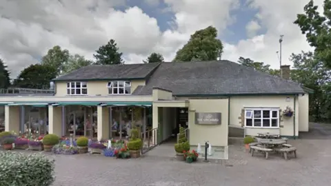 Street view image of the Italian Orchard in Broughton. A long cream rendered building with a grey roof and a white bow window to the right. To the left are three large glass window areas with many flowers and pots outside