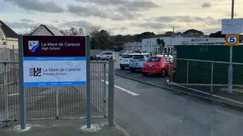 BBC The entrance to La Mare de Carteret school. showing metal gates. parked cares and signage. 