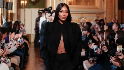 Getty Images Naomi Campbell walking in Torishéju's show at Paris Fashion Week in October 2023. Naomi is a 51-year-old black woman with her long hair perfectly straight. She wears a black shirt with exaggerated shoulders unbuttoned except for at the collar, exposing her midriff. She has her hands in the pockets of the black trousers it's paired with. The show is in a very grand room with wooden flooring and gold detailing. The runway is between audience members, many of whom are filming the collection on their smartphones