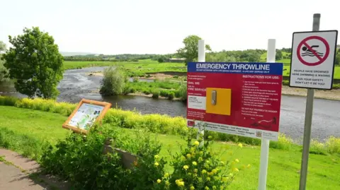 View from the banks of the River Ribble in Ribchester with signs detailing instructions in an emergency and a no swimming sign with a warning saying people have drowned in the river. 