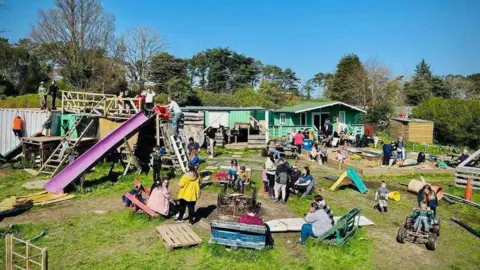 Isle of Play Children playing in the Lester's Yard playground, which features climbing frames made out of wooden ladders and pallets, and a purple slide. There is a green building in the background.