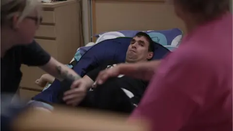 Stephen Fildes John Morrison lying down with carers around him