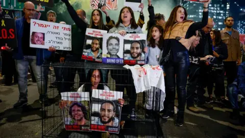 Reuters A woman sitting in a cage holds posters showing two Israeli hostages still being held by Hamas in Gaza, at a protest in Tel Aviv, Israel (26 March 2024)