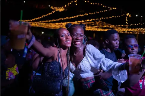 AFP Two young women smiling and posing for a picture with their drinks. It looks like night time and their are orange fairy lights behind them.