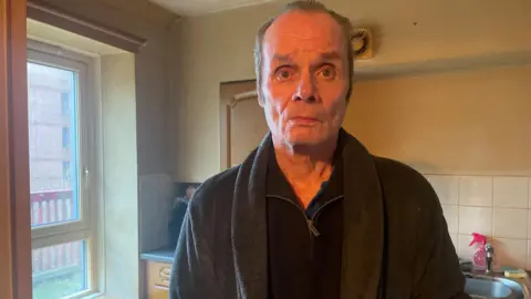 Simon Thake/BBC A man with thinning grey hair stands looking forlorn in a kitchen, wearing a dark dressing gown