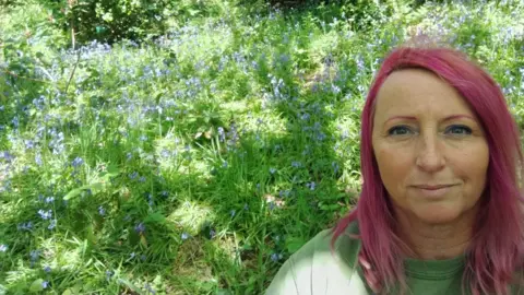Ayshea Cunniffe-Thomas Ayshea Cunniffe-Thomas, who has pink hair and pink eyebrows is stood in the woods, surrounded by bluebells on the grassy woodland floor. 