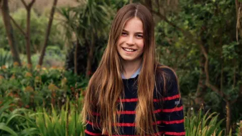 Photograph shows Charlotte wearing a black and red striped jumper and blue denim jeans, standing in a flowery field while smiling at the camera.