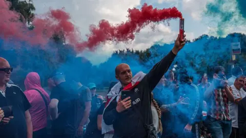 Getty Images A man holds a flare emitting red smoke during a protest outside the Bell Hotel in late July. He is wearing a long-sleeved dark blue top with the word 'air' written in white across the chest, and has an English flag tied around him like a cape. Around him are other protesters, and there is blue smoke in the air from similar flares.
