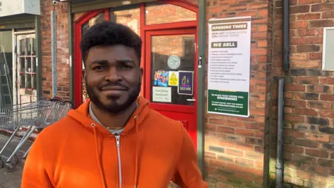 Photograph of Ifeanyi Chukwu, who runs the Peerless convenience store in Wigan town centre. The 36 year-old is pictured outside of his shop.