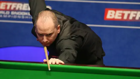 A balding man in a black shirt holds a snooker cue as he prepares to take a shot