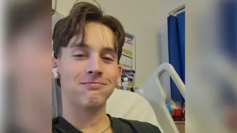 Contributed A picture of Ryan in hospital. He is taking a selfie style of photo and is smiling at the camera. He is lying in a hospital bed with headphones in his ears. He has brown, floppy hair that covers his forehead and he is wearing a black t-shirt.