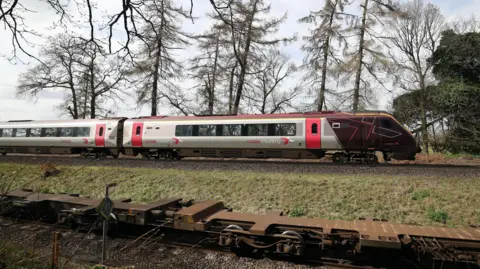 The cab and first carriage of a CrossCountry train travels on tracks in the countryside. In the background trees and fields can be seen.