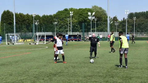 Two people in white shirts stand on a green football pitch, in the middle stands a referee in a black shirt, he is bald and pointing to his right. the ball is on the ground in front of him. to his left, stand two other players in yellow bibs