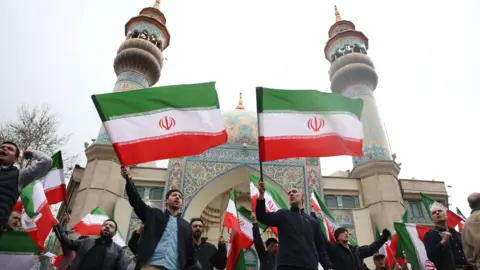 EPA A crowd of men wave Iranian flags outside an ornate mosque with two minarets.