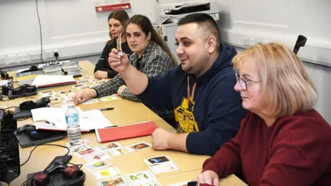 Hull City Council Four people are sat down at a table, there are three women and one man in the middle. The man is holding up a card and looking across the table and smiling. Laptops and mice devices can be seen on the other side of the table.