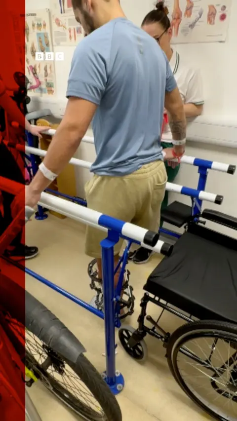 A man is stood and supporting himself by holding two rails with a wheelchair behind him