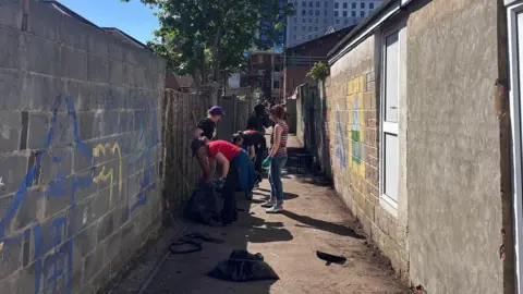 People in an alleyway with binbags and gloves, clearing rubbish from the floor. The alleyway has grey walls which have faded spray paint on them, and a dusty pathway down the middle.