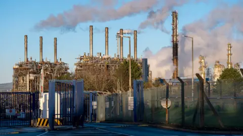 A general view of the ExxonMobil plant in Fife. There are several tall columns with dark smoke coming out of them and blowing into a blue sky.