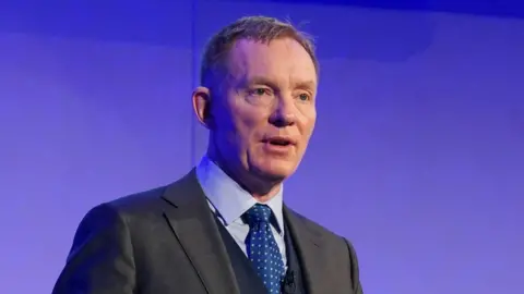 Getty Images Chris Bryant standing in front of a purple wall. He has a dark grey suit on and a light blue shirt, and blue tie. He is looking to the right of the camera and has short strawberry blonde hair. 