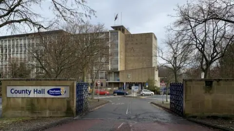 Six-storey building in glass and brick. It is viewed through a large set of open gates, on the left of which is a sign on the saying "County Hall" with the Durham County Council logo.