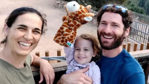 Maya Roman A photo of Yarden Roman-Gat, her husband Alon and three-year-old daughter Gefen, smiling as the child holds up a giraffe glove puppet