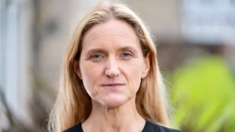 PA Media Labour MP Kim Leadbeater looks straight at the camera, in front of a blurred background. She has long hair, and is wearing a black top.