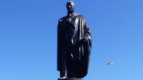 A large statue of a man standing, with a seagull flying past behind it.