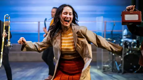 Will Green Photography Claire Lee Shenfield playing the role of Caroline in the Caroline musical. It is a production shot and the character is dancing to the radio. She is wearing a patterned mustard and white sweater with a burnt orange skirt and trench coat over the top.