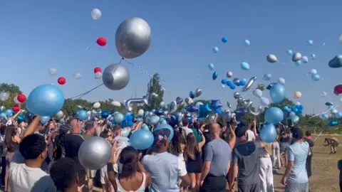 A crowd of people stood on grass against the backdrop of a blue sky with arms outstretched to release the balloons