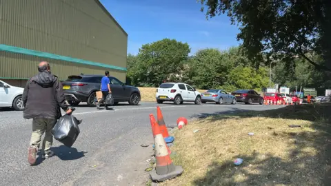 Aisha Iqbal/BBC A queue of cars waiting outside a recycling centre. The road leading up to it has grass verges and orange and white cones on the side. Two men on foot carry black bags of rubbish towards the gate.