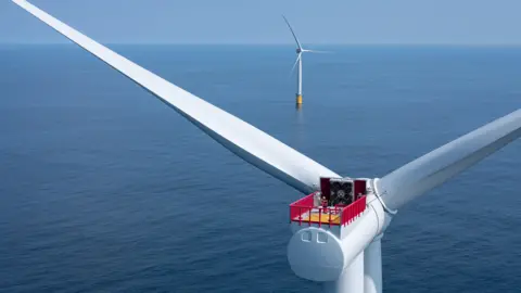 Reuters Two wind turbines at sea, one close to the camera showing only the turbine and part of the blades. The other is seen in the distance, with a yellow marking at the base of the white tower that supports the turbine. It is a clear day with calm waters.