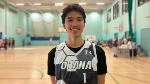 A young man looking into the camera and smiling. He has black hair and is wearing a basketball jersey which reads 'Ohana' on it. In the background, there is a basketball court with some people on it