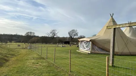 A pleasant day on farm land. There is a tee-pee and some animals in the background. There are clouds in the sky and some trees can be seen in the background. 