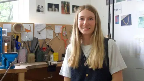 Iona smiles from her desk in an art studio. She has long, straight blonde hair and wears a black knitted vest over a white T-shirt. Behind her there are lots of tools such as chisels and saws, and postcards on the walls. 