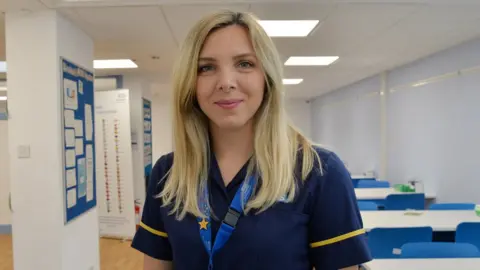 Ana-Maria Toth Ana-Maria Toth looks at the camera smiling. She is standing in a hospital ward wearing her nurses scrubs