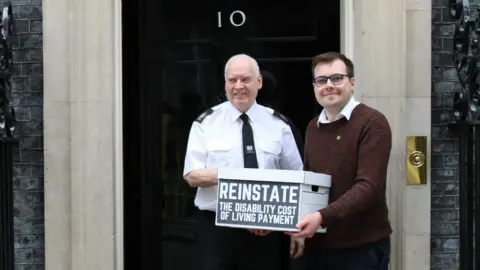 Supplied Tom Howard, on the right of the image, wearing a brown shirt over a white shirt while standing outside Downing Street holding a cardboard box with the word REINSTATE on it He is standing next to a security guard.