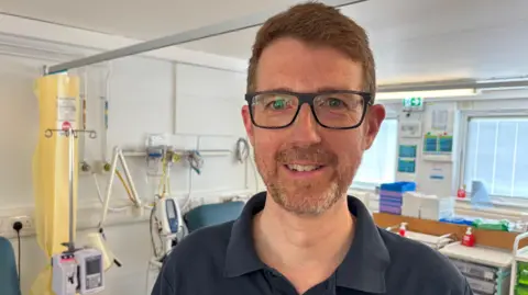 Dr Waters, who has short red hair and glasses, is wearing a blue t-shirt in a medical setting. Behind him are monitors, beds and a desk.