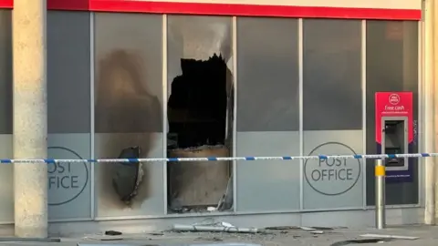 A close up of the post office on Midsummer Boulevard, Milton Keynes. It shows one of the ground-length windows has been smashed and a blackened interior. Police tape is rapped along the front of the building. 