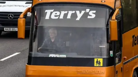 Lee Hall Terry Hall driving a orange Terry's Coaches coach on a busy road with a white Mercedes van in the lane next to him. A school sign is in the windscreen.