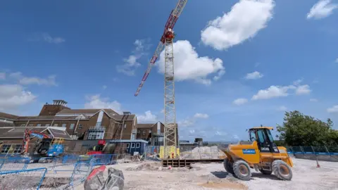 LDRS A large red and white crane in the middle of a building site, with a large brown-brick building to its right hand side.