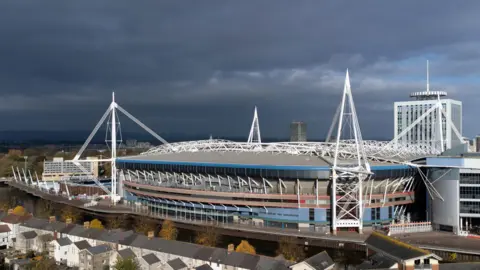 An aerial view of the Principality Stadium. Stormy grey skies are seen behind it, while in front runs the river and trees and a row of terraced houses. 