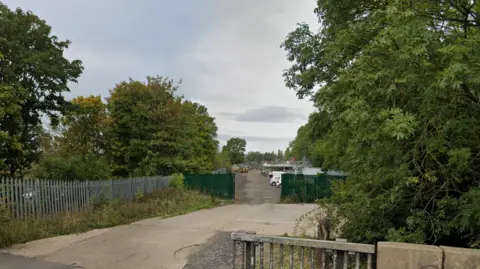 An industrial looking site behind green fences with trees surrounding the site