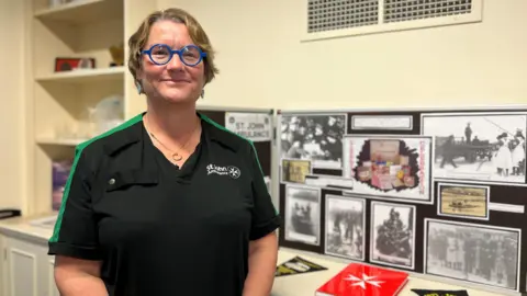 Emma smiles at the camera and she's wearing a St John Ambulance t-shirt and has glasses on with thick blue circular frames. Emma has short blonde hair and she's standing by a exhibition of St John Ambulance memorabilia 