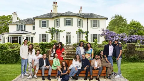 ITV/Amy Brammall The cast of the ITV dating show My Mum, Your Dad pose in front of the country house where the show is set. They are grouped in their pairs so children with the mum or dad. Four of the pairs are sat on the brown sofa which is on a large rug on a green grass lawn. The other four pairs are stood behind the sofa. Lots are leaning on the other person or have their arm around then. Behind them are some stone steps and bushes, and then a grand white country house.