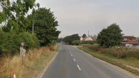 A grey rural road with white markings runs down the centre of the photo with verges of grass, shrubs and green trees to either side. A partially broken 40mph sign stands to the left. Houses can be seen in the background as the road curves to the left.