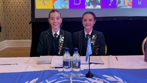 Two girls wearing dark green school uniforms and smiling at the camera. They both have dark hair tied back. On the table in front of them are some notes, two bottles of water and a flag of the United Nations.