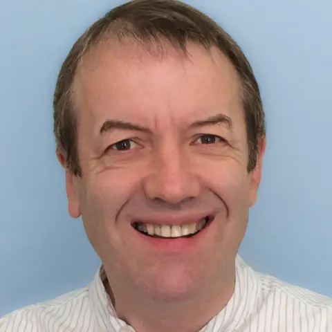 Dr Tony Collins A man in his 60s with brown hair is smiling broadly. He is wearing a white striped shirt.