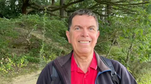 BBC/Sofía Luis-Hobbs A man in a red polo and navy jacket smiles at the camera in front of a woodland area.